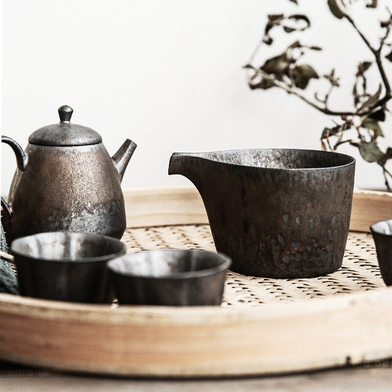 A vintage style ceramic creamer with long beak-shaped spout and a speckled black and gold finish, along with a matching teapot and cups, displayed on a bamboo serving tray.