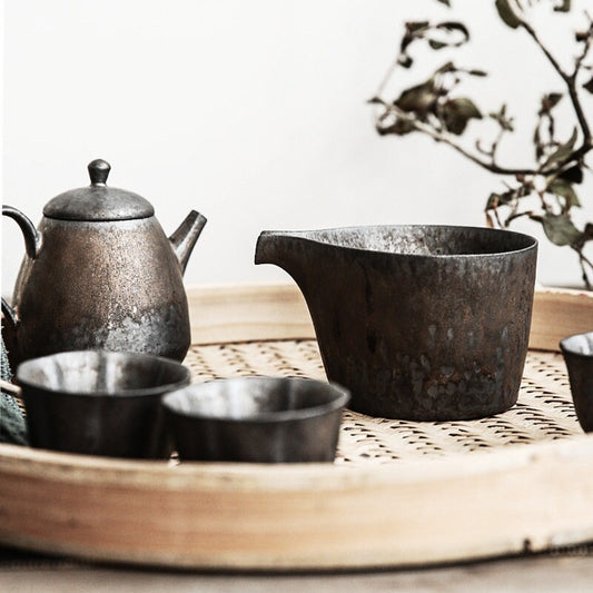 A vintage style ceramic creamer with long beak-shaped spout and a speckled black and gold finish, along with a matching teapot and cups, displayed on a bamboo serving tray. 